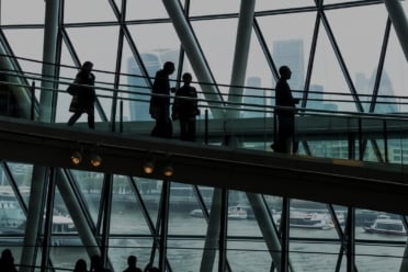 People walking on an elevated walkway inside a modern glass building with a cityscape and river visible through the windows in the background, echoing the atmosphere of insightful events like What I Heard at FORTRA: Why Your Lender Matters in Today’s Market.