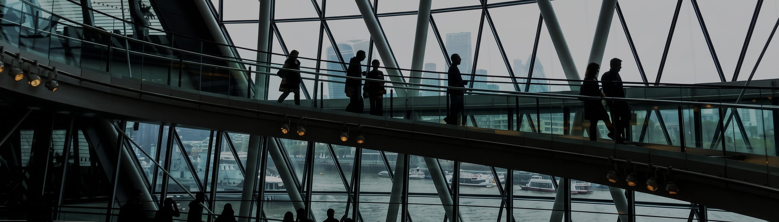 People walking on an elevated walkway inside a modern glass building with a cityscape and river visible through the windows in the background, echoing the atmosphere of insightful events like What I Heard at FORTRA: Why Your Lender Matters in Today’s Market.